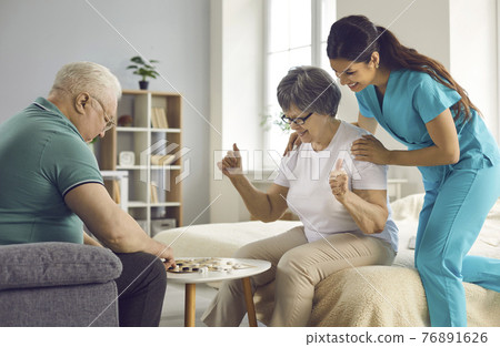 Supportive care giver watching couple of her happy senior patients play checkers Supportive care giver watching couple of her happy senior patients play checkers 76891626