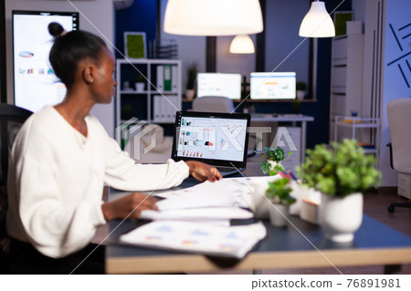 African woman working late at night looking at charst on laptop 76891981