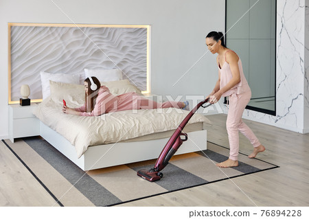 Using cordless vacuum cleaner to clean bedroom, mother does cleaning while daughter listens to music. 76894228