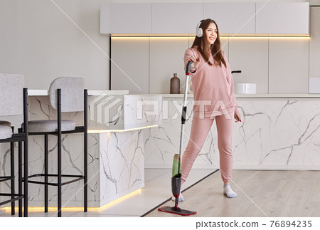 Using flat spray mop to clean kitchen, young woman in pink clothes tidies up and listens to music. Using flat spray mop to clean kitchen, young woman in pink clothes tidies up and listens to music. 76894235