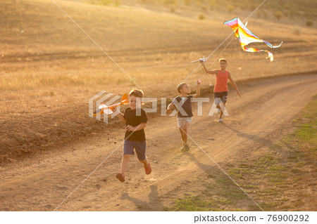 three joyful children, boys, run along a dirt road and launch toy airplanes and a kite, in backlight, concept 76900292