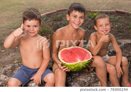 three happy children are sitting outdoors with a cut watermelon and smiling, two children show an affirmative gesture three happy children are sitting outdoors with a cut watermelon and smiling, two children show an affirmative gesture 76900294