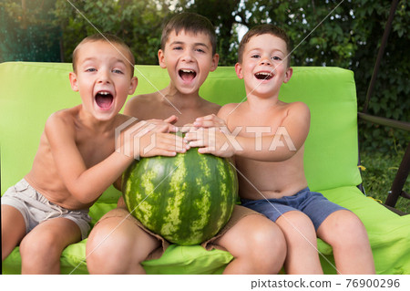 three boys are sitting on a bench, holding a big watermelon and laughing, summer concept 76900296
