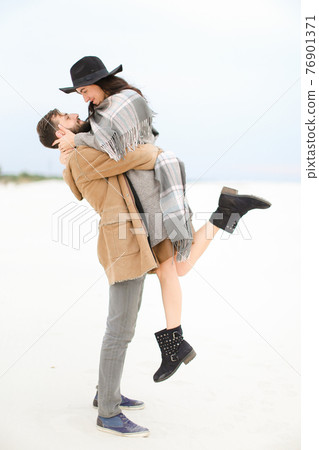 Man picking up young woman wearing hat, coat and grey scarf on white snow background. 76901371