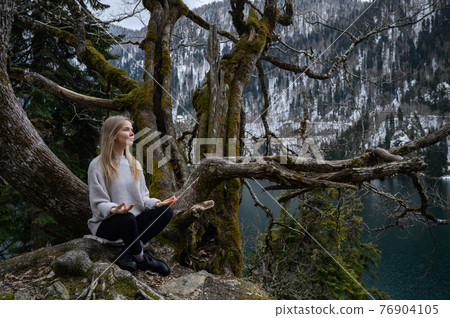 Girl sits on a tree and meditates on blue lake background. Lake Ritsa in Abkhazia 76904105