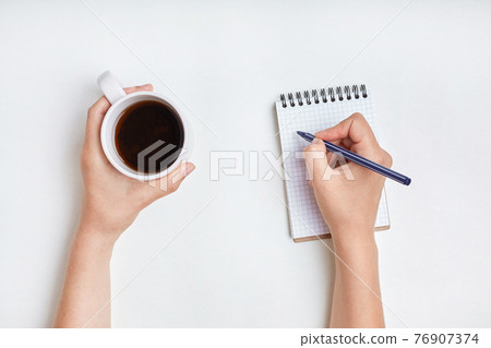 Top view of female hand writes in spiral notebook, takes notes with cup of tea. Woman writes in her diary. Beautiful female's hand holds pen above notebook paper with mug of coffee Top view of female hand writes in spiral notebook, takes notes with cup of tea. Woman writes in her diary. Beautiful female's hand holds pen above notebook paper with mug of coffee 76907374
