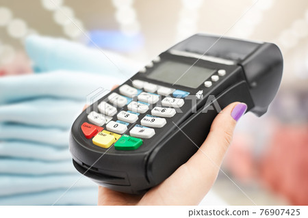 Indoor shot of woman`s hand holds POS terminal against boutique interior, pays with credit card for purchase. Credit card machine near pile of clothes 76907425