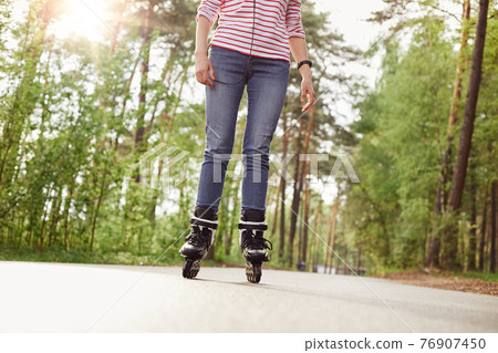 Faceless portrait of slim female wearing jeans and striped shirt, rollerblading on asphalt road surrounded with trees, spending time in forest in active way, unknown person roller skates outdoors. Faceless portrait of slim female wearing jeans and striped shirt, rollerblading on asphalt road surrounded with trees, spending time in forest in active way, unknown person roller skates outdoors. 76907450