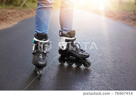 Horizontal picture of unknown person rollerskating, rollerskates being on wet road, having sun on background, black modern rollerskates used in everyday life, leisure activities. Movement concept. 76907454