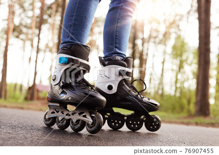 Close up picture of rollerblades, unknown person wearing rollerskates to spend time actively, black rollerblades with wheels being on road in fresh air, trying new purchase. Rollerskating concept. 76907455