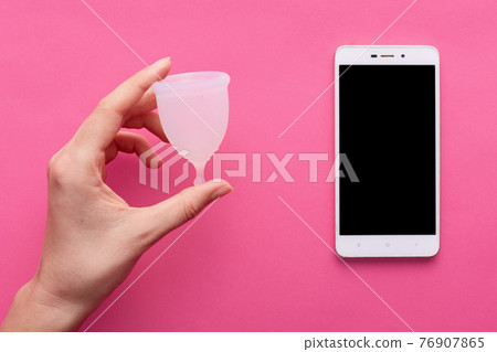 Flat lay of unknown hand holding rubber menstrual cup, smartphone with blank screen lying nearby isolated over pink background, app for menstruation periods, comfortable usage during periods. Flat lay of unknown hand holding rubber menstrual cup, smartphone with blank screen lying nearby isolated over pink background, app for menstruation periods, comfortable usage during periods. 76907865