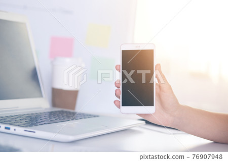 Picture of hand holding smartphone with blank screen, workplace with white laptop and papercup of coffee on desk, work at home using modern technologies, gadgets. People and freelance concept. Picture of hand holding smartphone with blank screen, workplace with white laptop and papercup of coffee on desk, work at home using modern technologies, gadgets. People and freelance concept. 76907948