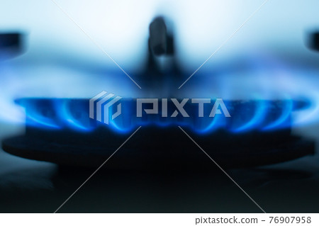 Close up picture of gas hob burning, bright blue flames situated around metallic special round in gas cooker, having light blue background, fire burning at minimum equally. Economy of gas concept. Close up picture of gas hob burning, bright blue flames situated around metallic special round in gas cooker, having light blue background, fire burning at minimum equally. Economy of gas concept. 76907958