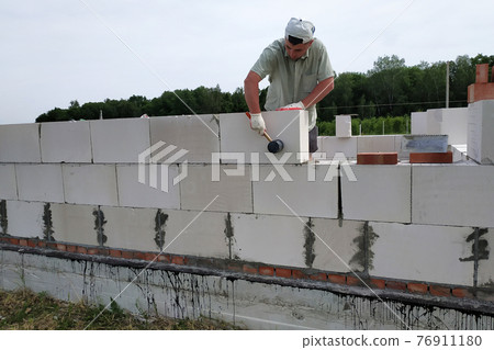 The master performs construction work, namely, laying a wall of gas blocks with tools and adhesive mortar, in the background the sky and the forest. The master performs construction work, namely, laying a wall of gas blocks with tools and adhesive mortar, in the background the sky and the forest. 76911180