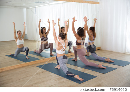 Group of young sporty women in yoga studio, practicing yoga pose in front of the mirror. Healthy active lifestyle, working out indoors in gym 76914030