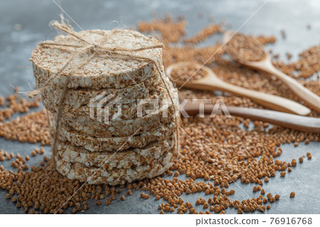 Spoons of raw buckwheat and delicious crispbread on marble surface 76916768