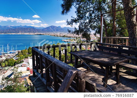 View of Fethiye harbor with yachts and boats and mountains. There are wooden table with benches for rest on foreground.  76916887