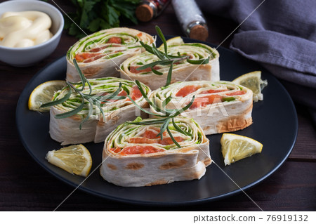 Rolls of thin pita bread and red salted salmon with lettuce leaves on a black ceramic plate, dark wooden background. 76919132