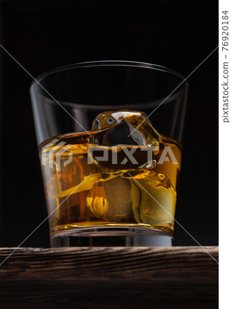 Glass of single malt whiskey with ice cubaes on top of wooden board on black. 76920184