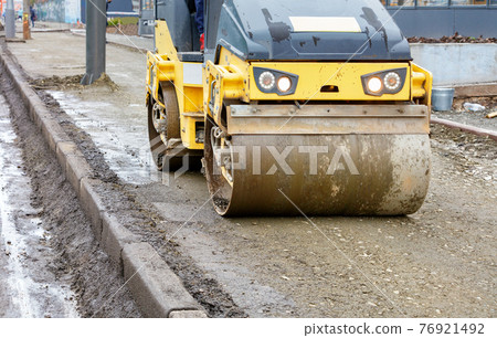 Frontal view of a road roller rolling the base of the sidewalk along a concrete curb near the carriageway. Frontal view of a road roller rolling the base of the sidewalk along a concrete curb near the carriageway. 76921492