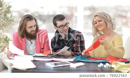 Smiling tailor businesswoman sitting at sewing machine and making handmade clothing while marketing assistant working on new projec Smiling tailor businesswoman sitting at sewing machine and making handmade clothing while marketing assistant working on new projec 76922563