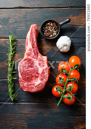 Raw uncooked black angus Bone In Steak in rustic style on wooden table background. with tomato garlic herbs and peppercorns rosemary close up Raw uncooked black angus Bone In Steak in rustic style on wooden table background. with tomato garlic herbs and peppercorns rosemary close up 76931860
