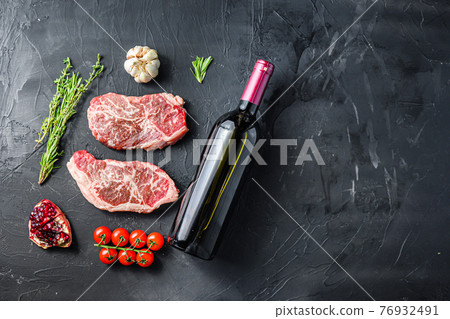 Organic top blade steak cuts, with red wine bottle and glasses, herbs and seasonings on black textured background top view with space for text. Organic top blade steak cuts, with red wine bottle and glasses, herbs and seasonings on black textured background top view with space for text. 76932491