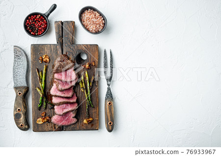Grilled fillet mignon beef steak, with onion and asparagus, on wooden serving board, with meat knife and fork, on white stone background, top view flat lay, with copy space for text 76933967