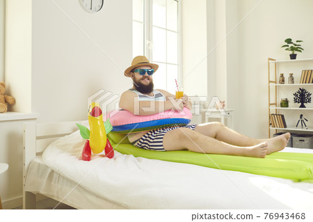 Funny bearded man with travel accessories lying in bed imagining resort vacation 76943468
