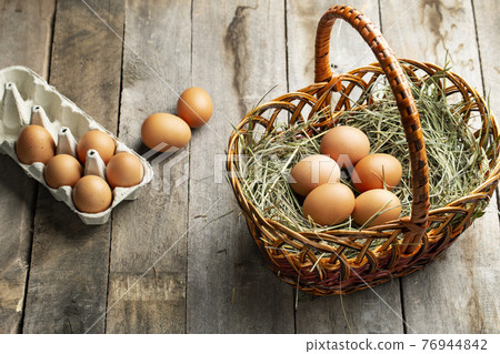 Chicken eggs in a hay basket and egg tray, on a wooden table. 76944842