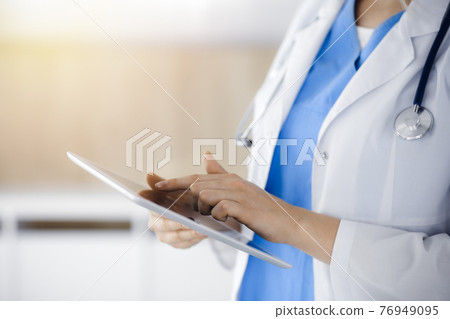 Unknown woman-doctor standing in sunny clinic and using tablet pc, close-up. Data and best service in medicine 76949095