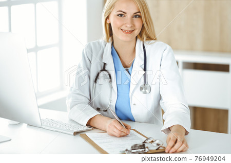 Portrate of woman-doctor at work while sitting at the desk in clinic. Blonde cheerful physician ready to help patients Portrate of woman-doctor at work while sitting at the desk in clinic. Blonde cheerful physician ready to help patients 76949204