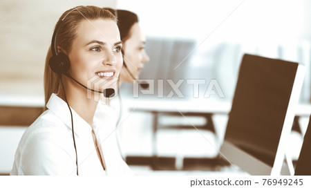 Blond female customer service representative and her colleague are consulting clients online using headset. Call center and business people concept 76949245