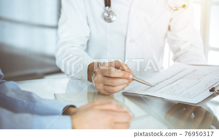 Unknown male doctor and woman-patient discussing current health examination while sitting in sunny clinic and using clipboard. Good medical service in hospital 76950317