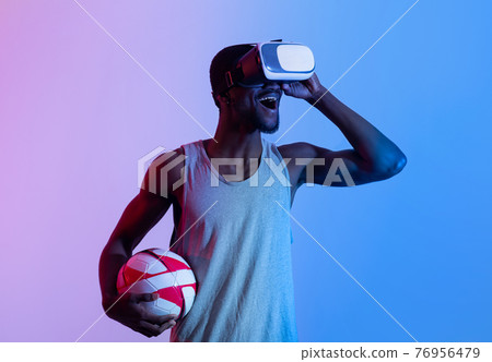 Exercising with virtual reality. Excited young black guy in VR headset holding soccer ball in neon lighting 76956479