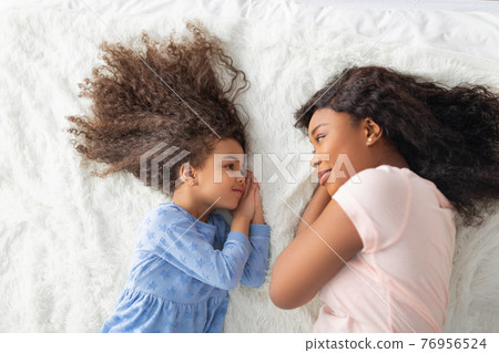 Little black girl with her mom in casual clothes lying on bed together, looking at each other, top view Little black girl with her mom in casual clothes lying on bed together, looking at each other, top view 76956524