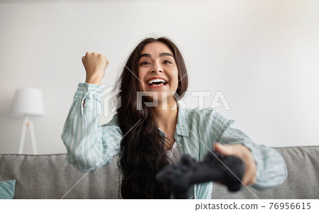 Happy Indian woman with controller playing online video game, making YES gesture at home Happy Indian woman with controller playing online video game, making YES gesture at home 76956615