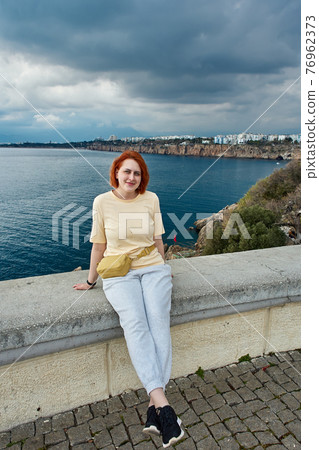 Young smiling redhead Caucasian woman posing for photographer while sitting on fence of observation point overlooking sea in Antalya. 76962373