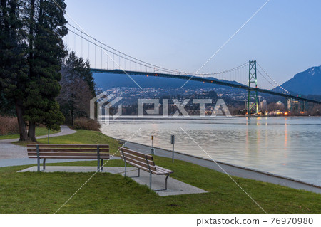 Stanley Park Seawall wooden bench. Lions Gate Bridge In the background at twilight. Vancouver, British Columbia, Canada. Stanley Park Seawall wooden bench. Lions Gate Bridge In the background at twilight. Vancouver, British Columbia, Canada. 76970980