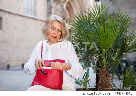 Elderly woman is looking for something in her handbag Elderly woman is looking for something in her handbag 76974943