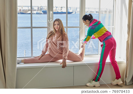 Mother helps her daughter, a cheerleader, to do stretching at home in order to be able to sit on twine during period of self-isolation and quarantine due to coronavirus. Mother helps her daughter, a cheerleader, to do stretching at home in order to be able to sit on twine during period of self-isolation and quarantine due to coronavirus. 76978433