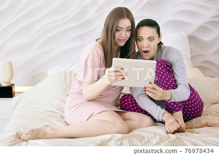 Online viewing of horror movie on tablet pc screen by two women sitting on bed in night clothes. Online viewing of horror movie on tablet pc screen by two women sitting on bed in night clothes. 76978434