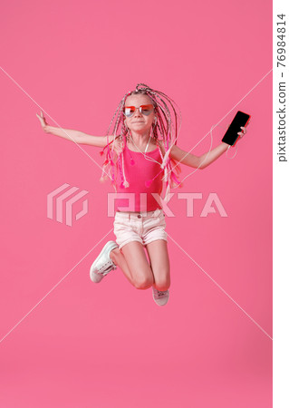 Beautiful little girl with dreadlocks jumping with mobile phone in the hand and listen music on headphones on pink background 76984814