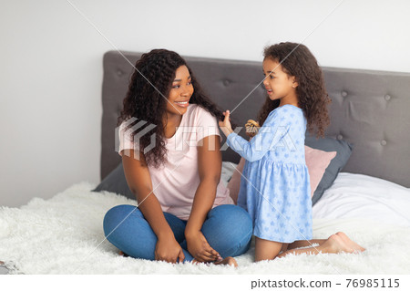 Adorable black girl brushing her mother's hair on bed indoors. Home spa salon concept Adorable black girl brushing her mother's hair on bed indoors. Home spa salon concept 76985115