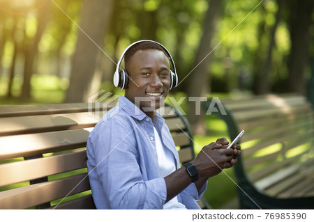 Handsome black man relaxing on bench in park and enjoying his favorite music on smartphone Handsome black man relaxing on bench in park and enjoying his favorite music on smartphone 76985390