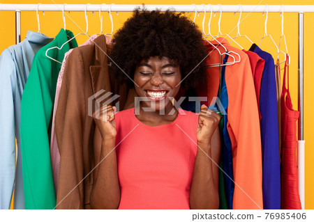 Excited Black Lady Shaking Fists Near Clothing Rail, Yellow Background 76985406