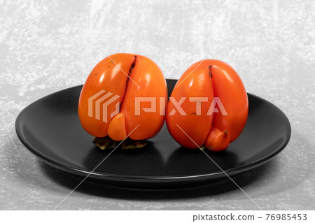 Two ugly ripe orange persimmons on black plate on light grey textured backdrop close up. Zero waste food concept. Reasonable sustainable consumption. Persimmon - source of vitamins and trace elements. 76985453