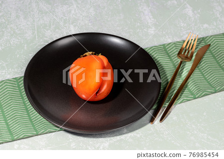 One ugly orange persimmon on black plate and golden cutlery on napkin on light backdrop. Zero waste food. Fresh healthy fruits. Reasonable sustainable consumption. Persimmon - source of vitamins. 76985454