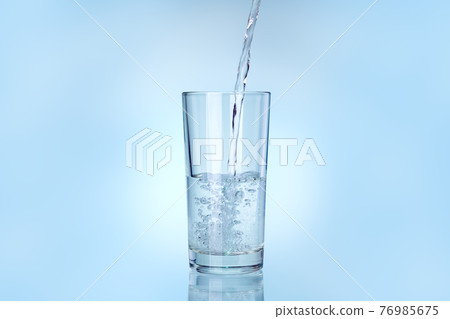 transparent glass is filled with water from a stream of liquid stream, drinkware on a blue background with copy space. 76985675