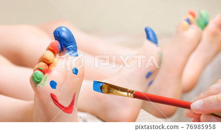 Close up hand brush drawing funny face on feet children with painted toes. Baby art therapy child painting face on legs kids paints smile on feet closeup. Sensory play drawing on feet kids fingers leg Close up hand brush drawing funny face on feet children with painted toes. Baby art therapy child painting face on legs kids paints smile on feet closeup. Sensory play drawing on feet kids fingers leg 76985958